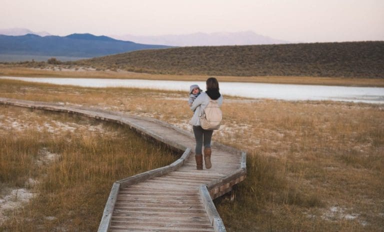 Maman qui se promène avec son enfant en portage, sur un chemin de bois bordant la mer, lumière rasante